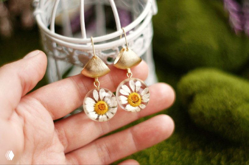 Пара подвесных серёжек с небольшими ромашками, зажатыми в прозрачной смоле, держат в руке на размытом зелёном фоне.