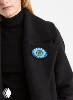 Брошь в виде глаза с ромашкой в центре, прикреплена к тёплому тёмному пальто; сочетание аксессуара и одежды в образе.