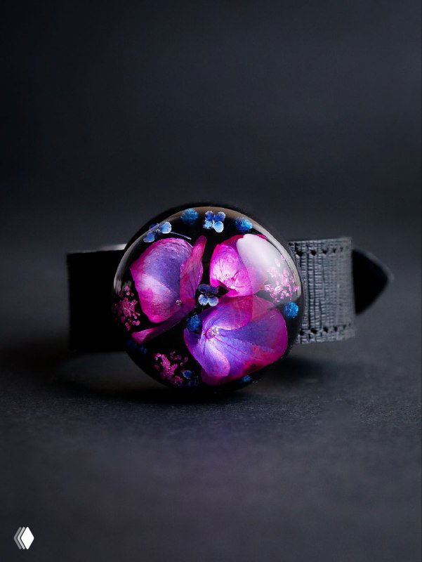 Маленькая круглая брошь-кабошон с розово-фиолетовыми лепестками гортензии в прозрачной смоле, показана на матовом чёрном фоне.