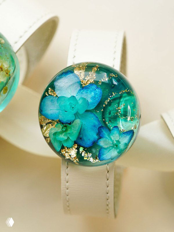 Круглая бусина с бирюзово-зелёными засушенными гортензиями и золотыми вкраплениями в прозрачной смоле на светлом фоне, идеальна для браслета или кулона.