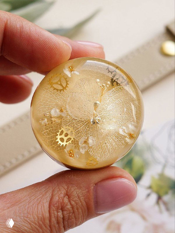 Круглый элемент с белыми и золотистыми деталями: фрагмент гортензии в прозрачной смоле, показан крупным планом в руке мастера для масштаба.