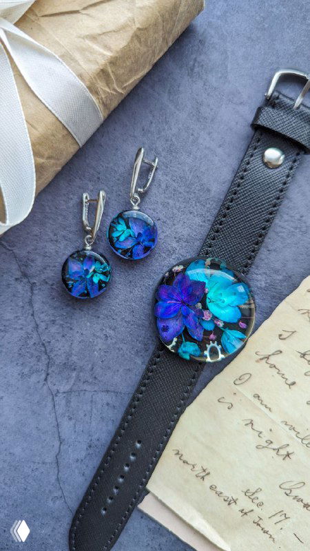 Набор из украшений: колье и серьги с синими и фиолетовыми частями гортензии в смоле, размещённые на ремешке часов и декоре для фотосъёмки.