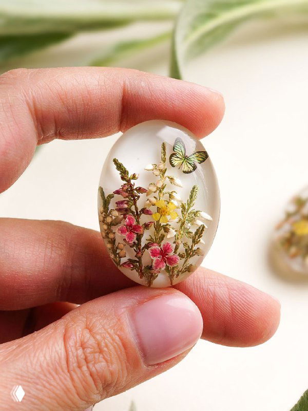 Овальный кабошон с небольшими полевыми цветами и алиссумом в прозрачной смоле, изображение демонстрирует масштаб изделия в руке