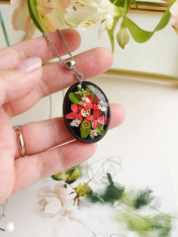 Крупный план кулона с застывшими в смоле мелкими цветами и зеленью, украшение держат пальцы на светлом фоне домашней студии.