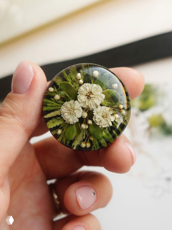 Круглая брошь в руке с букетиком белых соцветий и папоротниковых листьев, чёткая фактура просушенной зелени в эпоксидной заливке.