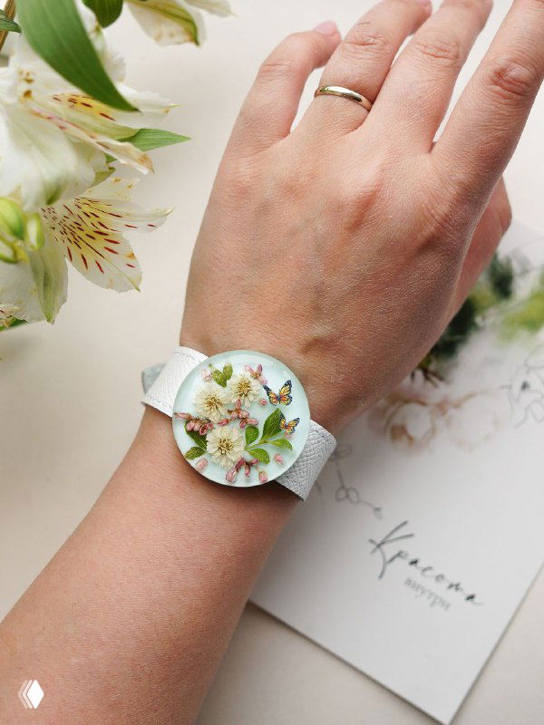 Белый ремешок часов и крупный кабошон с зелёными листьями и мягкими цветочными акцентами на запястье в повседневном сеттинге.