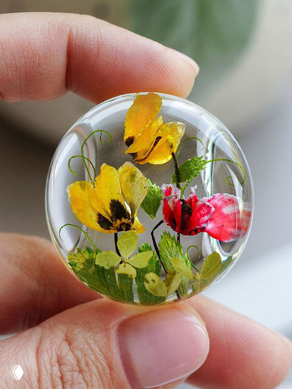 Крупный план прозрачного стеклянного кулона с яркими жёлтыми и красными цветами и зелёными стеблями, композиция выглядит натуральной и свежей.