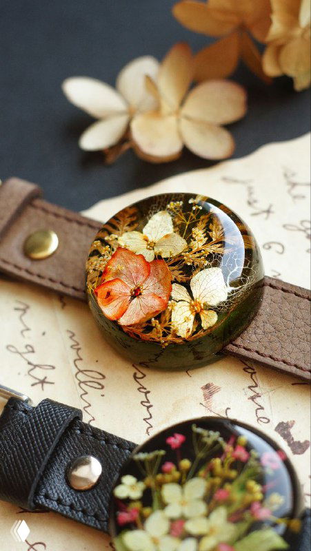 Крупный план круглых украшений из прозрачной смолы с аккуратно уложенными засушенными цветами и мелкой зеленью, на фоне бумаги и ремешков.