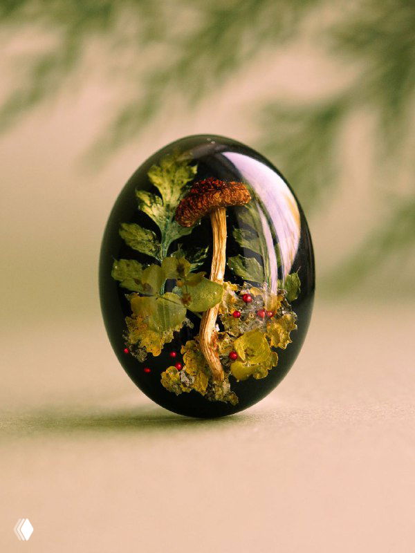 Овальная кабошон-подвеска из смолы с миниатюрной сушёной грибной композицией и зелёной листвой внутри на нейтральном фоне.