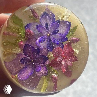 Круглый кабошон с пурпурными и розовыми цветами в прозрачной смоле; показана глубина, блеск поверхности и внутренняя текстура материала.