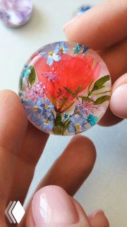 Крупный план круглого прозрачного кабошона из смолы с засушенными цветами — герань и незабудки, изделие держат пальцы для демонстрации размера и текстуры.