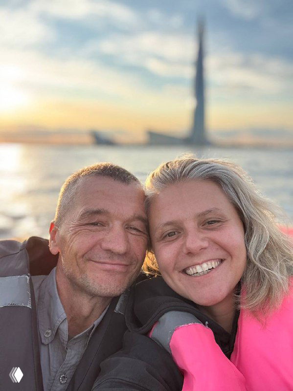 Два взрослых человека на селфи у воды на закате в спасательных жилетах, улыбаются в камеру; на фоне видны силуэты городской застройки и море.