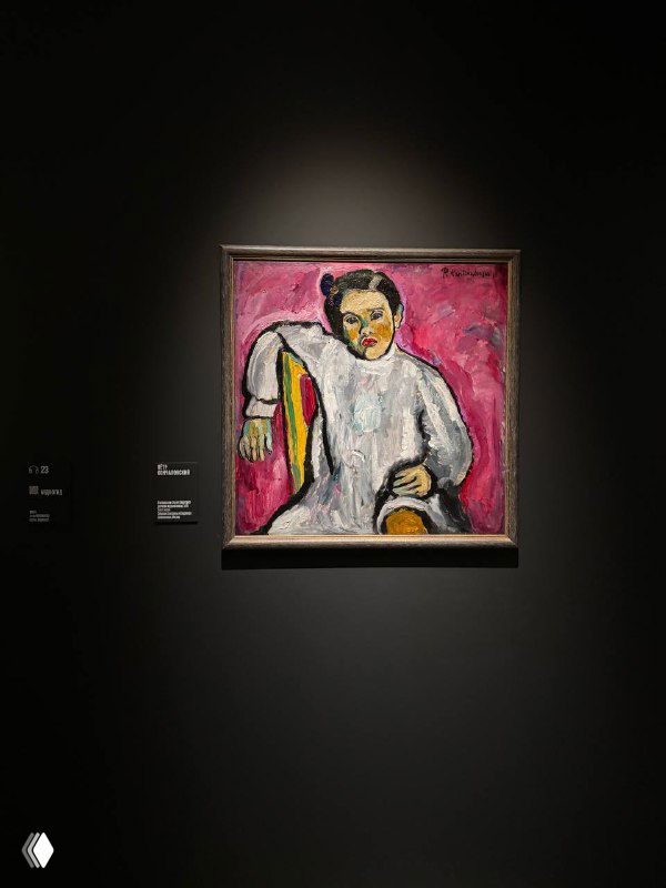 Картина в раме на тёмном стенде из выставки: портрет с розовым фоном, заметна текстура кисти и приглушённое музейное освещение.