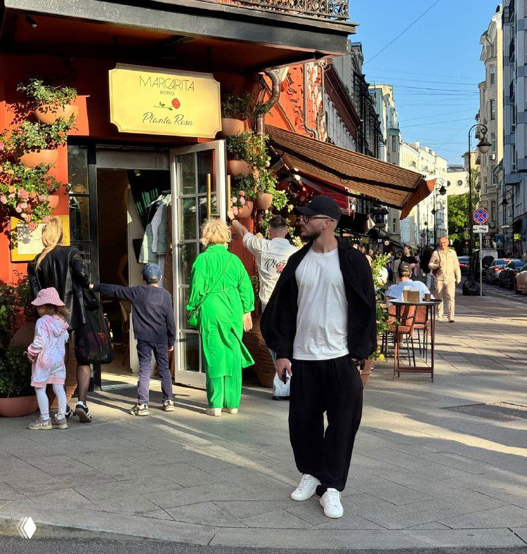 Городская уличная сцена: мужчина в чёрной толстовке и белых кроссовках идёт мимо кафе, на тротуаре прохожие, одна женщина в ярком зелёном наряде и летняя атмосфера улицы.