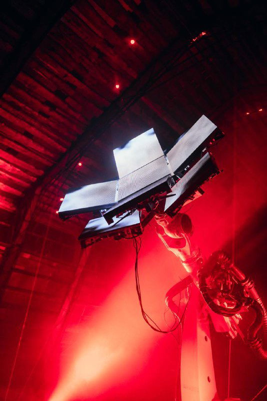 Деталь механической конструкции инсталляции на насыщенно-красном фоне: кабели, крепления и источники света подчёркивают технологическую эстетику проекта.