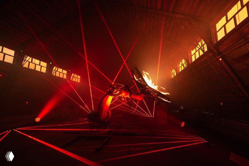 Роботизированная рука и платформа в большом пространстве с красным освещением и лазерными линиями, подчёркивающими масштаб перформанса и инженерную работу команды.