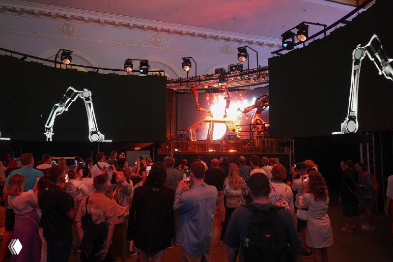Публика на мероприятии смотрит сцену с проекциями и роботизированными манипуляторами; сочетание технологий, музыки и перформанса создает атмосферу шоу.
