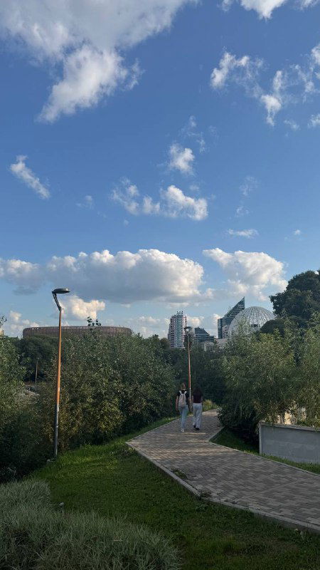 Аллея в городском парке Екатеринбурга с зелёными насаждениями и широким небом; дорожка уходит в перспективу, видны пешеходы и городская архитектура.