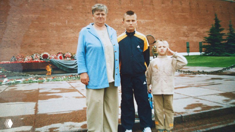 Цветное фото на фоне мемориала: пожилая женщина, мужчина в спортивной форме и мальчик, семейный снимок у памятника с цветами.