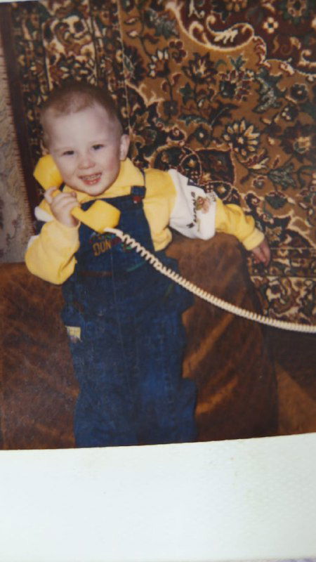 Фотография малыша в комбинезоне, держащего телефонную трубку на фоне узорного ковра — домашний снимок из раннего детства.