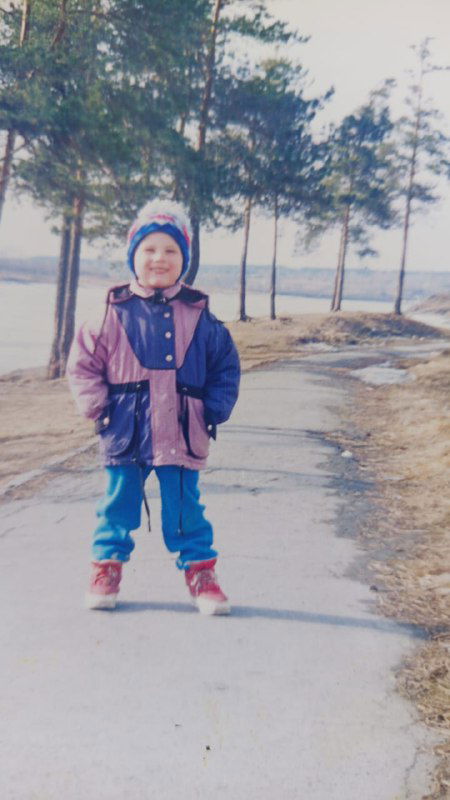 Ребёнок в тёплой куртке и шапке идёт по дорожке вдоль берега и соснового леса — кадр прогулки в провинции или пригороде.