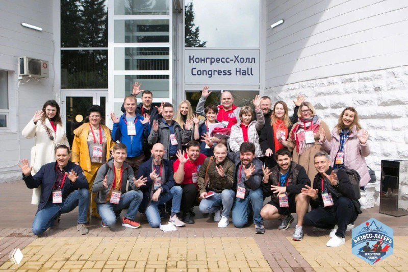 Групповое фото участников бизнес-лагеря у входа в конгресс-холл: люди с бейджами, улыбки и командная атмосфера.