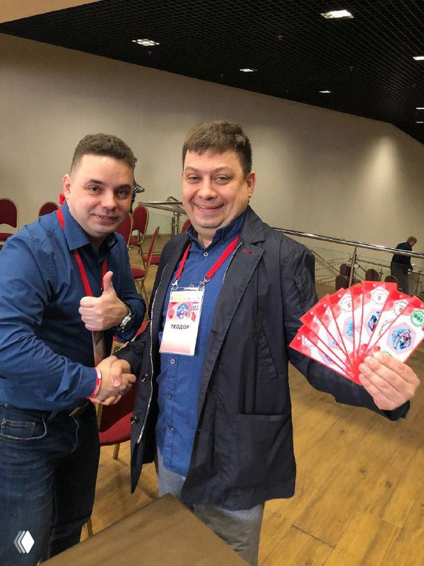 Фотография двух участников/организаторов мероприятия в зале: люди в бейджах обмениваются рукопожатием и демонстрируют набор купонов или призов на мероприятии