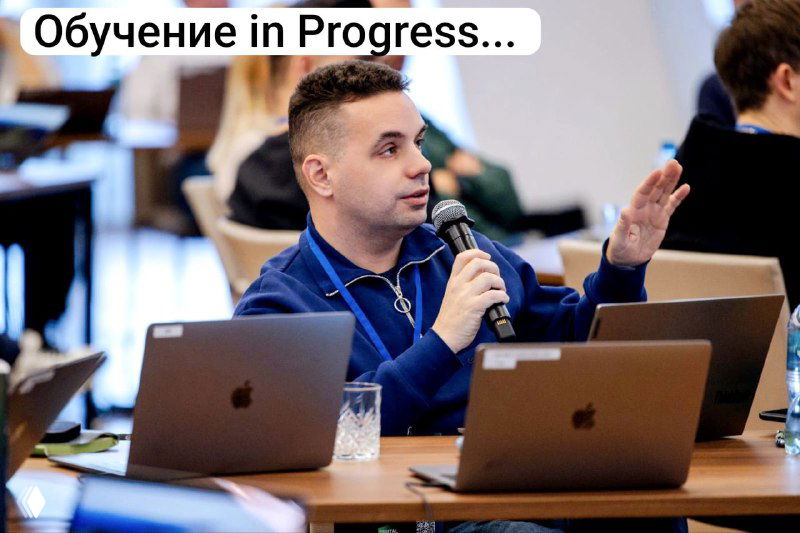 Фото: мужчина говорит в микрофон за столом с ноутбуками — сцена обучения или выступления на профессиональном мероприятии.