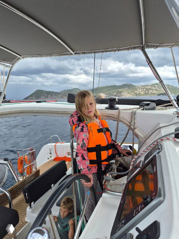Девочка в спасжилете стоит в кокпите парусной яхты, вокруг элементы палубы и снастей, спокойное море и берег на фоне, атмосферный кадр семейного путешествия.