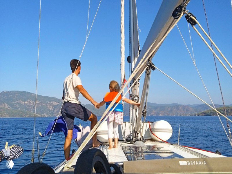 Взрослый ведёт ребёнка за руку по носовой палубе парусной яхты, спокойное море и прибрежные горы на горизонте, семейная прогулка под парусом в хорошую погоду.