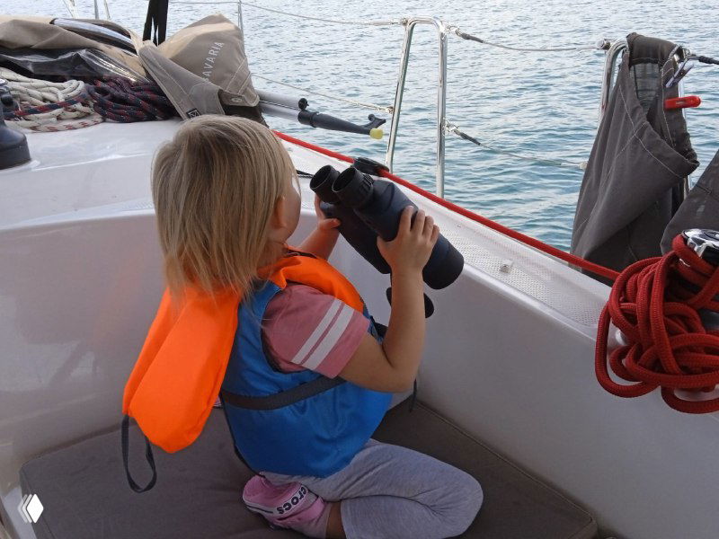 Ребёнок в спасательном жилете сидит в кокпите и рассматривает берег через бинокль, рядом канаты и элементы палубы — типичная сцена семейного похода на яхте.