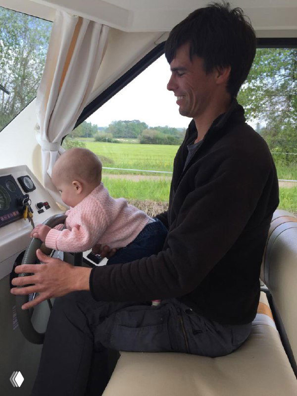 Отец за штурвалом моторной лодки держит на коленях маленькую дочь, внутри видна уютная каюта и окна с видом на зелёный берег, семейная атмосфера на борту.