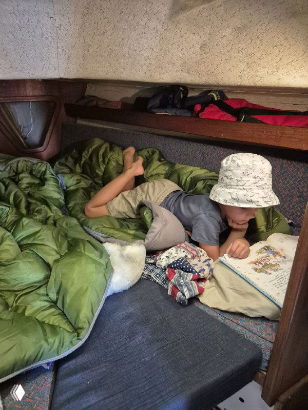 Ребёнок в спальнике в носовой каюте яхты читает книгу, вокруг уютное внутреннее пространство, вещи экипировки и тёплая атмосфера семейного плавания ночью или в тихой стоянке.