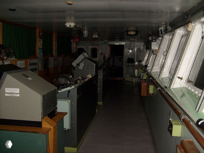 Интерьер капитанского мостика с навигационными пультами, приборами и окнами, приглушённое освещение в рабочей рубке.