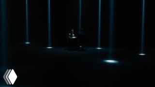Тёмный сценический кадр из рекламного ролика: одиночная фигура за столом в центре, вокруг направленные вертикальные световые лучи и атмосферное освещение.