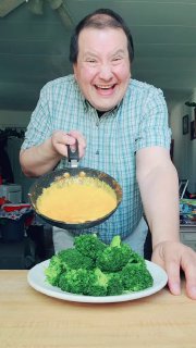 Человек с улыбкой держит сковороду с яичной смесью над тарелкой с брокколи — кадр похож на копию формата «странная еда + танец» в тренде TikTok.