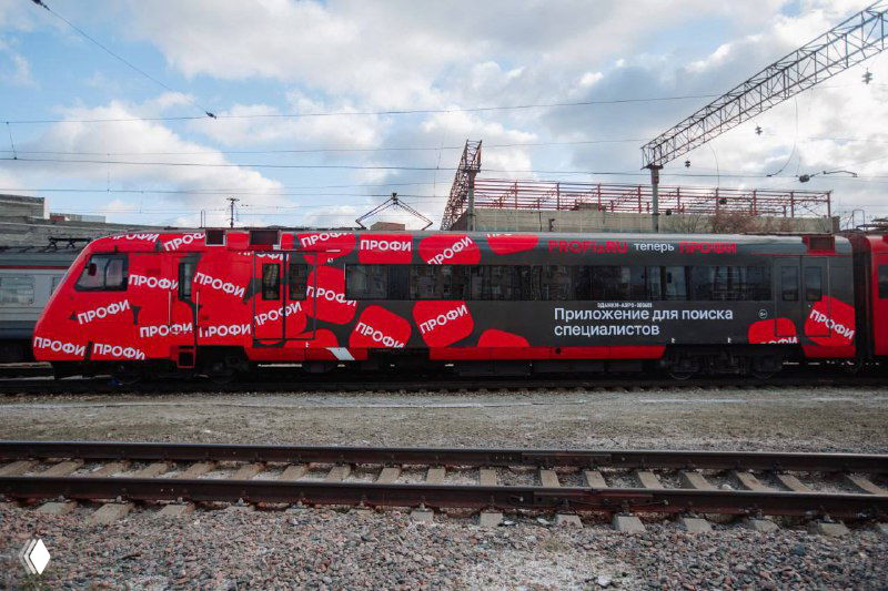 Поезд Аэроэкспресса, оклеенный красочным брендингом «Профи», стоит на железнодорожной платформе под открытым небом в рамках наружной кампании.