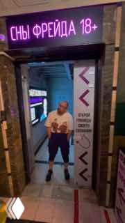 Неоновая вывеска «СНЫ ФРЕЙДА 18+» над входом в помещение, в дверном проёме стоит мужчина в шортах и футболке; ночной городской кадр, передающий атмосферу уличного снимка в контексте обсуждения вирусных видео.