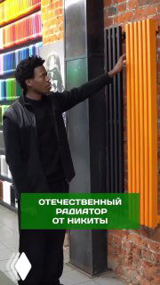 Человек в тёмной одежде прикасается к оранжевому радиатору на фоне кирпичной стены и цветных элементов интерьера; кадр из обсуждаемого ролика и подкаста.