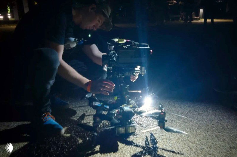 Техник в ночной съёмке на коленях у сложного рига с освещением на земле, демонстрирующий масштаб подготовки и использование дополнительного света.