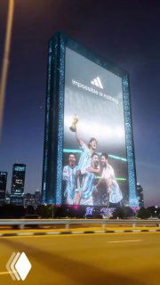 Ночной кадр фасада небоскрёба с огромным билбордом: 3D-изображение Месси с трофеем и логотипом бренда, снято с дороги при уличном освещении.