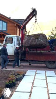 Грузовик с краном разгружает большой валун во дворе частного дома: рабочие направляют крепление и снимают камень с платформы.