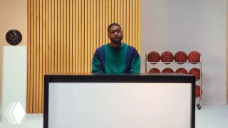 Кадр рекламного ролика: мужчина стоит за белым прилавком на фоне декоративной деревянной стенки и ряда баскетбольных мячей; студийный эстети́ческий кадр.