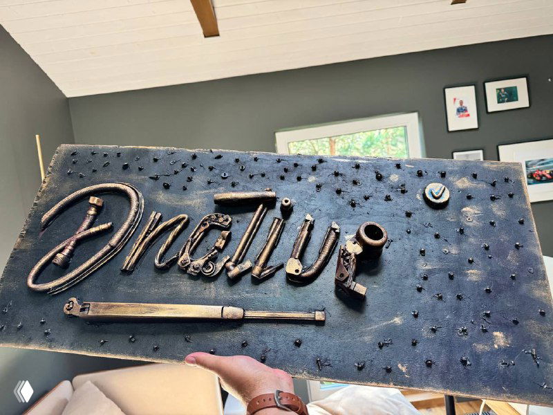 Деревянное панно с металлизованным логотипом «Dnative» в руках человека на фоне домашнего интерьера; видны декоративные гвозди и рука держателя.