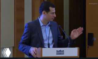 Мужчина в деловом костюме за трибуной, выступает на мероприятии — кадр из видео, иллюстрирующего обсуждаемую инициативу о соцрекламе.