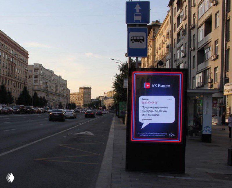 Ситилайт на городской улице с рекламой «VK Видео»: плакат в рамке с логотипом VK и диалоговым облачком, намёк на замедление YouTube, городской фон.