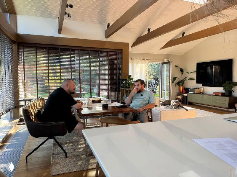 Два человека разговаривают за столом в светлой домашней студии: видна мебель, жалюзи на окнах и телевизор на заднем плане.