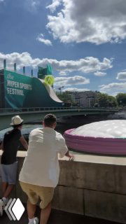 Двое людей смотрят на большую розово-белую надувную конструкцию у реки; на заднем плане облачное небо и городские здания.