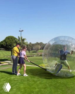 Игроки на поле: женщина готовится ударить по мячу перед прозрачной надувной сферой, в которой стоит человек; солнечный день и зелёное поле.