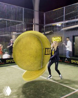 Ночной кадр паддл-корта: крупный желтый теннисный мяч крупным планом и играющие люди на фоне ограждений и рекламных баннеров.